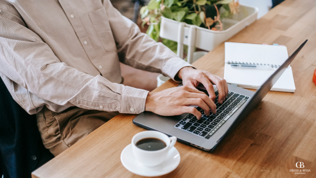 Homem trabalhando em um laptop em uma mesa de madeira, com uma xícara de café e material de escrita ao lado, em um ambiente iluminado e confortável com plantas decorativas.